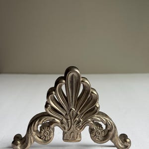 May include: A silver-toned, ornate metal decorative stand. The stand features a central fan-shaped design with floral accents and curved, scrolled legs. The decorative piece is likely for display.