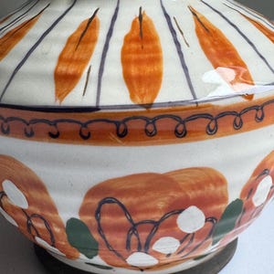 May include: A ceramic vase with a wide base and a narrow neck. The vase is painted with orange leaf-like shapes and blue stripes on a white background. The base is dark brown, and the rim has a red interior.