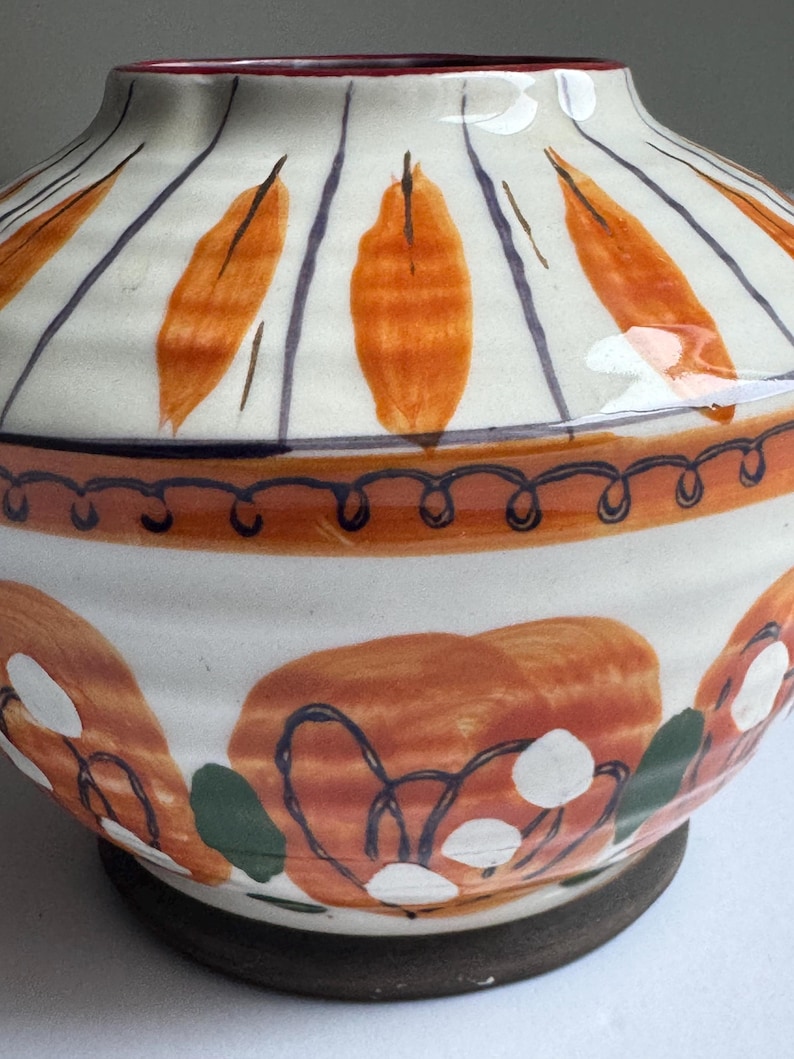 May include: A ceramic vase with a wide base and a narrow neck. The vase is painted with orange leaf-like shapes and blue stripes on a white background. The base is dark brown, and the rim has a red interior.