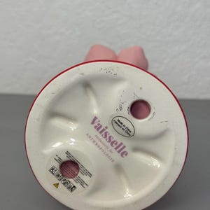 May include: A pink ceramic candle holder with a red rim and a bow-shaped top. The base is white and features the text "Vaisselle exclusively for ANTHROPOLOGIE". Made in China.