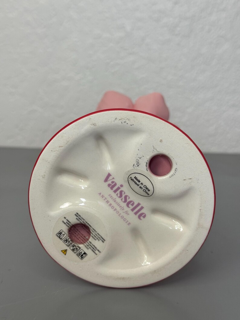 May include: A pink ceramic candle holder with a red rim and a bow-shaped top. The base is white and features the text "Vaisselle exclusively for ANTHROPOLOGIE". Made in China.