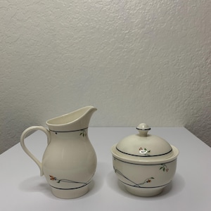 May include: Cream-colored ceramic creamer and sugar bowl set. The creamer has a handle and spout, while the sugar bowl has a lid with a small knob. Both pieces are decorated with a black line and floral accents.