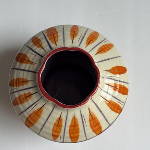 May include: A ceramic vase with a wide, round shape. The exterior is cream-coloured with orange leaf-like designs and blue vertical lines. The rim is red, and the interior is dark. The vase is likely handmade.