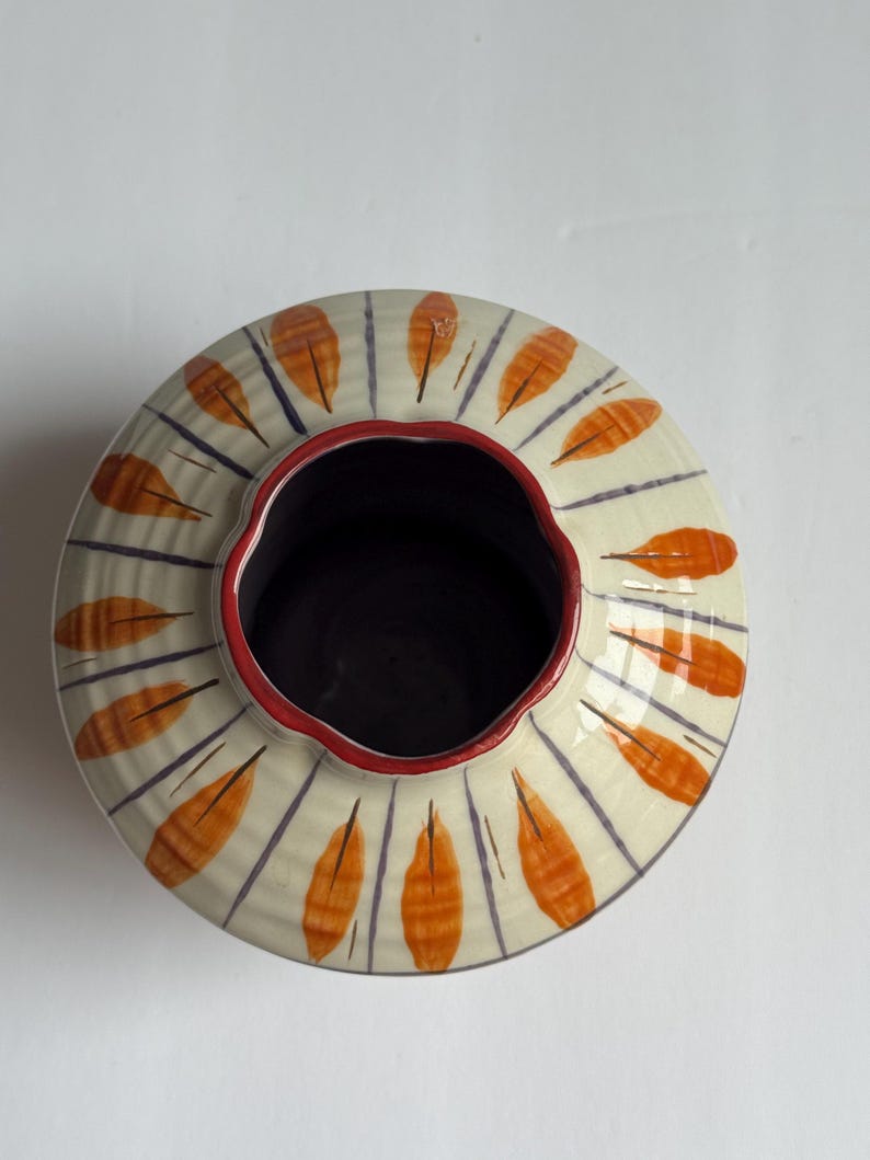 May include: A ceramic vase with a wide, round shape. The exterior is cream-coloured with orange leaf-like designs and blue vertical lines. The rim is red, and the interior is dark. The vase is likely handmade.