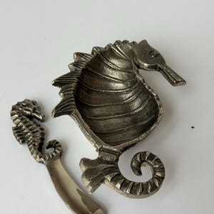 Può includere: Un piatto a forma di cavalluccio marino color argento con un cucchiaino abbinato. Il piatto ha una superficie strutturata e una grande ciotola aperta. Il cucchiaino ha un manico curvo. Il design del cavalluccio marino è dettagliato e decorativo.