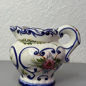 May include: A small, white ceramic creamer with a handle and spout. The creamer is decorated with blue, green, and pink floral designs and blue scrollwork. The base and rim are trimmed with blue. The creamer is approximately 10 cm tall.