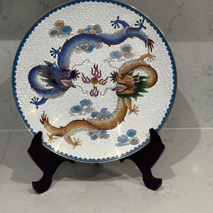 Puede incluir: Plato decorativo con dos dragones azules y dorados sobre fondo blanco con diseño. Borde azul y soporte de madera oscura. Los dragones están rodeados de motivos de nubes y llamas coloridas.