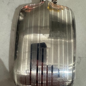 May include: A rectangular silver-toned metal flask featuring a vertical pinstripe pattern across the body. The flask has a rounded top with a screw-on cap and a central rectangular engraved frame on the front surface, reflecting light.