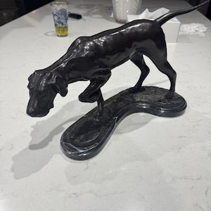 Op de afbeelding: Een donkerbronzen hondensculptuur in een jachthouding. De hond is gebeeldhouwd met gedetailleerde kenmerken, waaronder een lange staart en puntige oren. De sculptuur is gemonteerd op een donkere, gebogen basis.