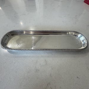 Könnte beinhalten: Ein langes, schmales, rechteckiges silberfarbenes Tablett mit abgerundeten Ecken und einem dekorativen Rand in Flecht- oder Seiloptik. Die Oberfläche des Tabletts ist glatt und reflektierend und weist durchgehend ein poliertes Metallfinish auf.