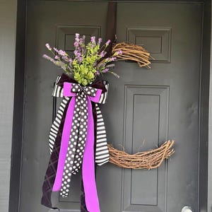 May include: A decorative grapevine wreath with a black and white striped bow, polka dot ribbon, and purple ribbon. The wreath is adorned with purple flowers and green foliage, hanging on a black door.
