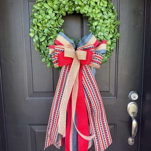 May include: A circular green boxwood wreath adorned with a large decorative bow. The bow features layered ribbons in red, white, and blue, including strips of burlap, solid red fabric, and blue fabric with white stars and red and white stripes.