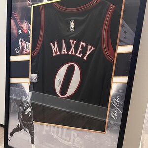 May include: Framed black basketball jersey with the name "MAXEY" and the number "0" in white. The jersey is black with red and white accents. The frame includes images of the player and the text "Tyrese Maxey".