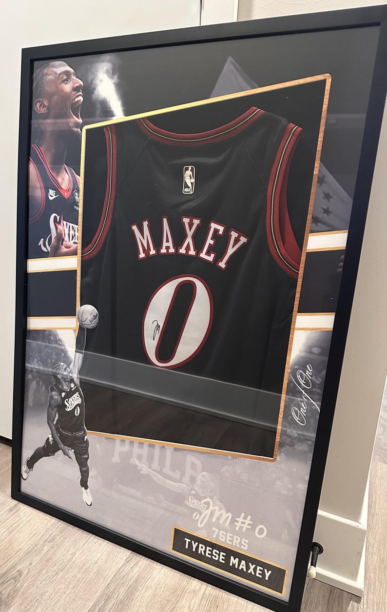 May include: Framed black basketball jersey with the name "MAXEY" and the number "0" in white. The jersey is black with red and white accents. The frame includes images of the player and the text "Tyrese Maxey".