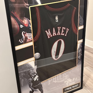 May include: Framed basketball jersey of Tyrese Maxey, number 0, in black with red trim. The frame includes images of the player in action and the text "PHILADELPHIA 76ERS". The jersey is signed.