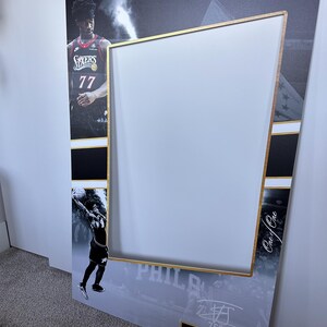 May include: A large photo booth prop featuring a basketball player in a 76ers jersey. The frame has a gold border and a white center for taking photos. The prop includes the text "76ERS" and "VJ EDGECOMBE".