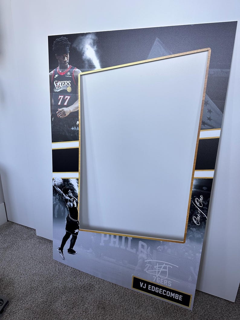 May include: A large photo booth prop featuring a basketball player in a 76ers jersey. The frame has a gold border and a white center for taking photos. The prop includes the text "76ERS" and "VJ EDGECOMBE".