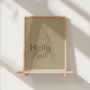May include: A framed print with the words "Hello Fall" in a decorative font. The print has a light brown background and is displayed on a small wooden shelf. The frame is made of light-colored wood.