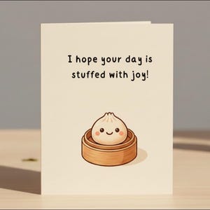 May include: A greeting card with the text "I hope your day is stuffed with joy!" features a cartoon dumpling in a bamboo steamer. A cupcake with pink frosting and a lit candle sits to the right. The card and cupcake are on a wooden surface.