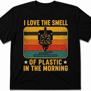 May include: A black short-sleeved t-shirt featuring a graphic of a 3D printer nozzle. The design includes a retro-style sunset background in yellow, orange, and red, with the text I LOVE THE SMELL OF PLASTIC IN THE MORNING printed in yellow.