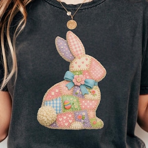 May include: Dark gray t-shirt featuring a patchwork bunny design. The bunny is composed of various pastel-colored fabric patches, including pink polka dots, gingham, and floral patterns. A light blue bow and a fluffy pom-pom tail complete the design.
