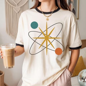 May include: A cream-colored ringer t-shirt with black trim on the collar and sleeves. The front features a graphic design of a gold starburst centered within black elliptical orbits, accompanied by a teal circle and an orange circle.