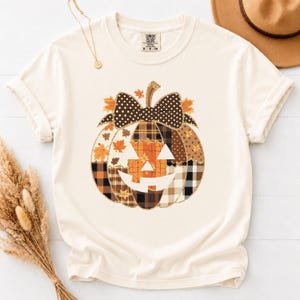 May include: A cream-coloured short-sleeve t-shirt featuring a large pumpkin graphic on the front. The pumpkin is composed of various brown, orange, and black plaid and leaf patterns, topped with a dark brown polka-dot bow and a jack-o'-lantern face.