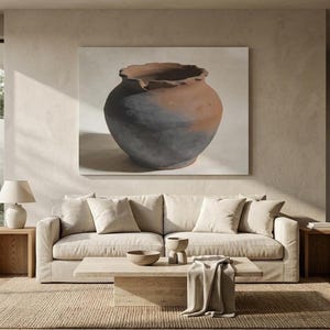 May include: A large rectangular wall art print featuring a round, wide-mouthed ceramic vessel with a textured, dark charcoal and terracotta finish, displayed above a beige sofa with matching cushions and a light-colored stone coffee table.