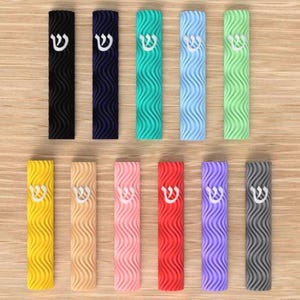 Könnte beinhalten: Eine Sammlung farbenfroher Mesusa-Gehäuse in verschiedenen Farbtönen, darunter Schwarz, Blau, Grün, Gelb, Rosa, Rot, Lila und Grau. Jedes Gehäuse hat ein strukturiertes Wellenmuster und einen weißen hebräischen Buchstaben. Die Gehäuse sind auf einer hellen Holzoberfläche angeordnet.