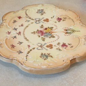 May include: A decorative, scalloped-edge ceramic plate with a floral design. The plate features a cream-coloured background with pink, blue, and yellow roses, accented with green leaves and blue ribbons. The edges are a soft peach colour with gold trim.