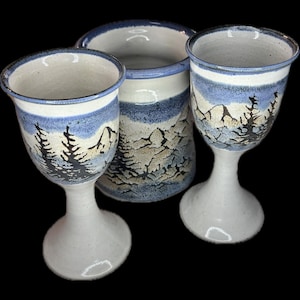 Puede incluir: Conjunto de tres piezas de cerámica: dos copas con pie y un jarrón cilíndrico. Cada pieza presenta un diseño de paisaje de montañas y árboles en azul, gris y negro sobre un fondo blanco. Las copas y el jarrón son decorativos.