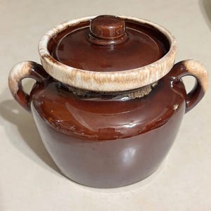 May include: A brown ceramic crock pot with a lid. The crock pot has two handles and a white and brown rim around the top. The lid has a small knob on top.