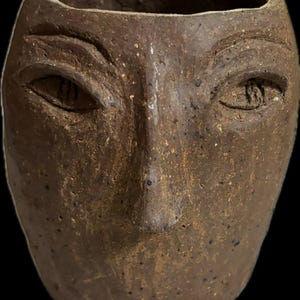 Peut inclure: Un pot de fleurs en céramique marron en forme de visage avec des traits sculptés, dont des yeux, un nez et une bouche. Le pot a une surface texturée et une large ouverture en haut. Une pièce de décoration intérieure unique.