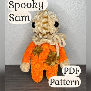 Puede incluir: Una muñeca de ganchillo con cabeza beige, ojos de botón negros y boca cosida. El cuerpo es de color naranja brillante con parches de color marrón oscuro y lleva un volante beige en el cuello. El texto Spooky Sam y PDF Pattern es visible.