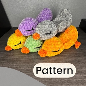 May include: A collection of six small, plush, crocheted duck toys made from soft, fuzzy yarn in yellow, purple, light green, gray, and orange. Each duck features a bright orange beak and closed eyes stitched in black thread. The word Pattern is visible.