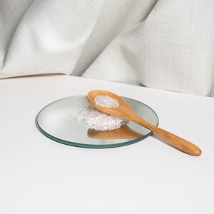 May include: A light brown wooden spoon rests on a small circular mirror. A small pile of translucent, pale pink crystalline salt grains sits on the mirror surface, partially underneath the bowl of the wooden spoon, reflecting the light.