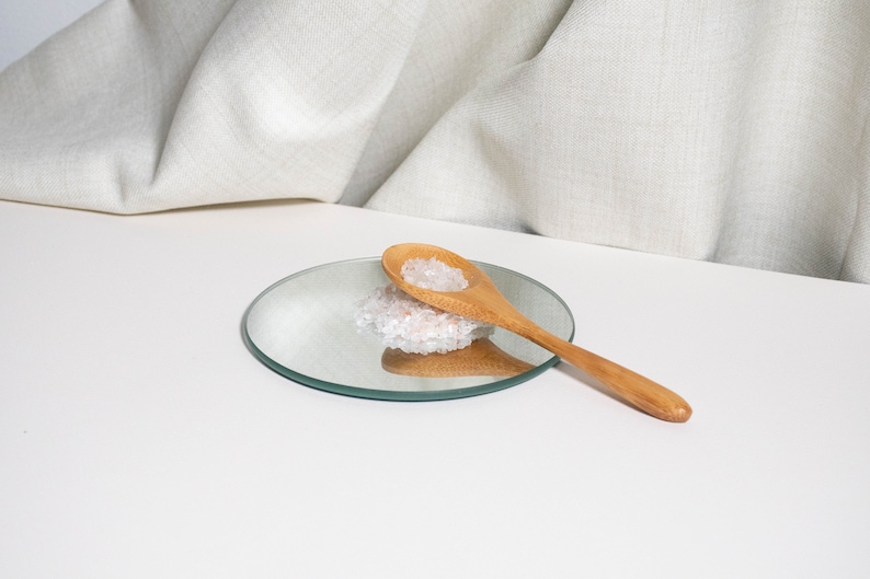 May include: A light brown wooden spoon rests on a small circular mirror. A small pile of translucent, pale pink crystalline salt grains sits on the mirror surface, partially underneath the bowl of the wooden spoon, reflecting the light.