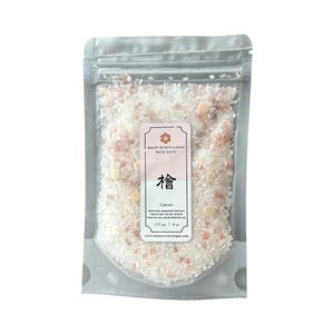 May include: A clear resealable pouch filled with white and pink bath salts mixed with small wooden cubes. The label reads Beauty Secrets of Japan Bath Salts, Cypress, with ingredients including Epsom salt, Himalayan pink salt, and 113 grams or 4 ounces.