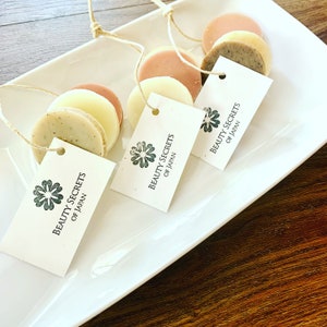 Puede incluir: Tres jabones redondos con etiquetas que dicen "Beauty Secrets of Japan" en un plato blanco. Los jabones están apilados uno encima del otro y están atados con una cuerda.