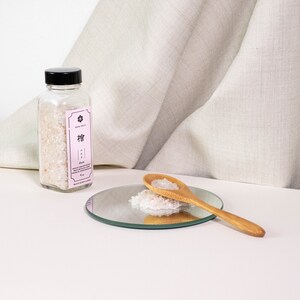 May include: A clear glass bottle with a black cap containing translucent crystals, featuring a light pink label with the text "檜" and "Hinoki". A wooden spoon rests on a small circular mirror, holding a pile of the same translucent crystals.