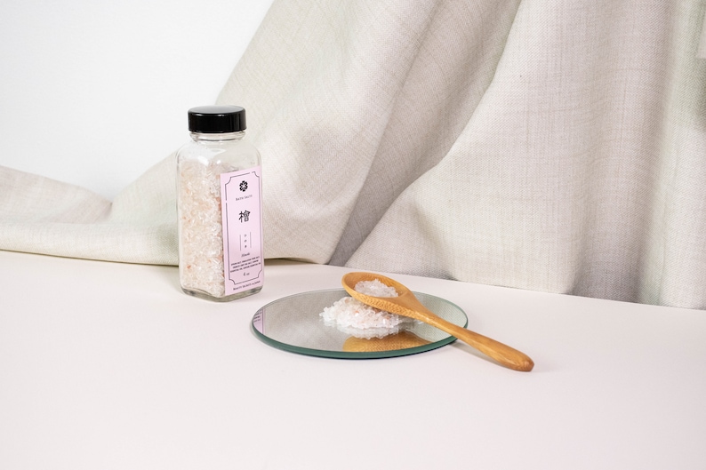 May include: A clear glass bottle with a black cap containing translucent crystals, featuring a light pink label with the text "檜" and "Hinoki". A wooden spoon rests on a small circular mirror, holding a pile of the same translucent crystals.