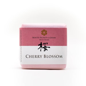 Könnte beinhalten: Eine rosa Badebombe mit einem weißen und weinroten Etikett. Auf dem Etikett steht "BEAUTY SECRETS OF JAPAN BATH BOMB CHERRY BLOSSOM" mit japanischen Schriftzeichen und einem floralen Design. Die Badebombe ist in rosa Seidenpapier verpackt.