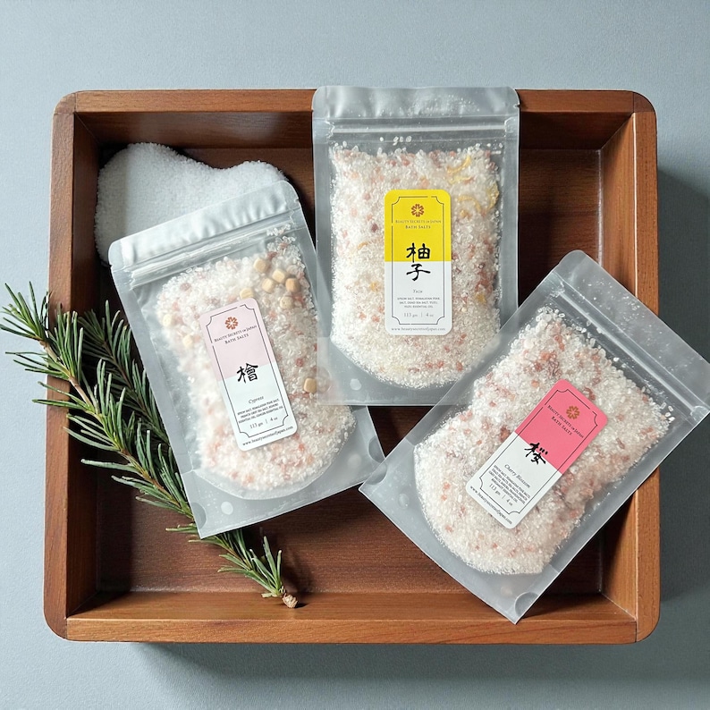 May include: Three clear resealable pouches filled with bath salts, each weighing 113 grams or 4 ounces. The labels feature the Japanese characters for Cypress, Yuzu, and Cherry Blossom. A green pine sprig rests inside a wooden tray with the bath salt pouches.