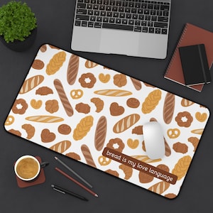 May include: A rectangular mousepad with a white background and a pattern of various baked goods in shades of brown and yellow. The phrase "bread is my love language" is printed on the mousepad. A computer mouse, laptop, and coffee cup are also visible.