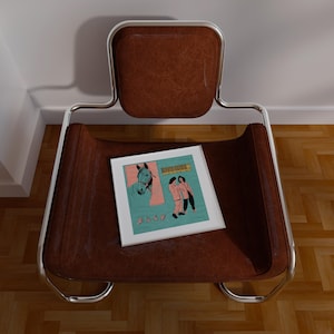 May include: A brown leather chair with a chrome frame. A framed print with a horse illustration and figures is on the seat. The chair is positioned against a white wall and a wooden floor.