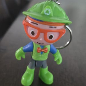 May include: A plastic keychain figure of a character wearing a lime green hard hat with a white recycling symbol, orange glasses, a blue shirt, a lime green vest, an orange bowtie, and lime green boots, attached to a silver metal keyring.