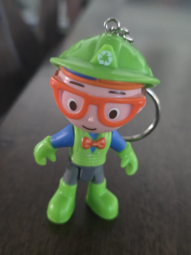 May include: A plastic keychain figure of a character wearing a lime green hard hat with a white recycling symbol, orange glasses, a blue shirt, a lime green vest, an orange bowtie, and lime green boots, attached to a silver metal keyring.