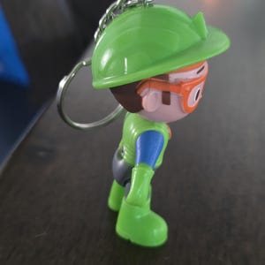 May include: A keychain featuring a small plastic figurine of a character wearing a bright green hard hat, orange safety goggles, a green vest over a blue shirt, and green boots. The figure has brown hair and is attached to a silver metal ring.