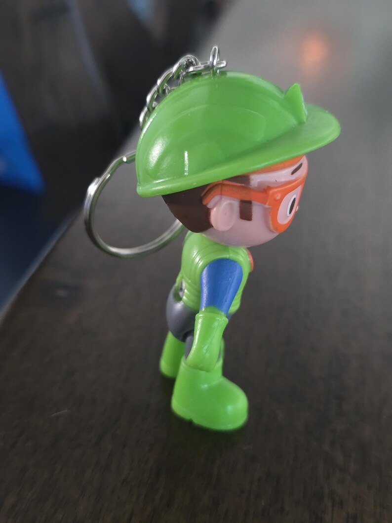 May include: A keychain featuring a small plastic figurine of a character wearing a bright green hard hat, orange safety goggles, a green vest over a blue shirt, and green boots. The figure has brown hair and is attached to a silver metal ring.