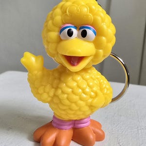 Może przedstawiać: Żółty brelok Big Bird ze srebrnym kółkiem. Figurka ma teksturowany korpus, pomarańczowe stopy i różowe opaski na nogach. Oczy są białe z niebieskimi i różowymi akcentami. Dziób jest otwarty, a ptak macha.